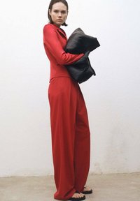 Costum roșu croit, cu mâneci lungi, pantaloni largi și o geantă oversized de tip pernă, neagră. Textură netedă, design minimalist și siluetă modernă.