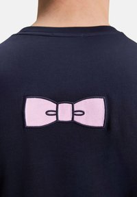 T-shirt en coton bleu marine avec un nœud rose brodé dans le dos, entouré de coutures noires. Texture douce, encolure ras du cou.