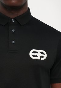 Közelkép egy fekete poloingről, három gombbal és egy fehér hímzett logóval a bal mellen.