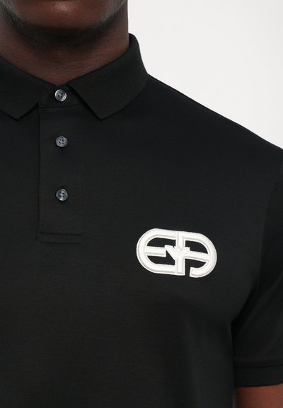 Prim-plan al unei cămăși polo negre cu trei nasturi și un logo brodat alb pe pieptul stâng.