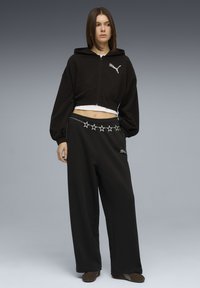 Νεαρή γυναίκα που φοράει μαύρο κοντό φούτερ με κουκούλα και μαύρο παντελόνι φαρδιάς γραμμής με ζώνη αλυσίδα σε σχήμα αστέρι, στέκεται μπροστά σε γκρι φόντο με διαβάθμιση.