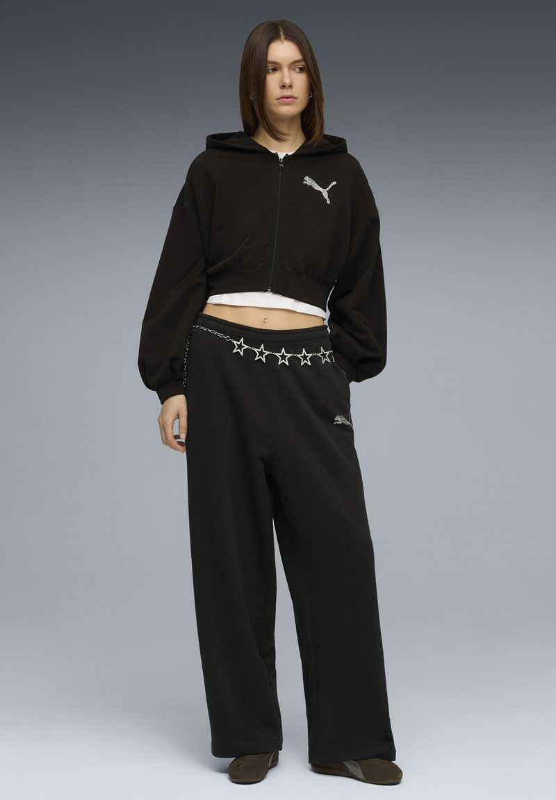 Νεαρή γυναίκα που φοράει μαύρο κοντό φούτερ με κουκούλα και μαύρο παντελόνι φαρδιάς γραμμής με ζώνη αλυσίδα σε σχήμα αστέρι, στέκεται μπροστά σε γκρι φόντο με διαβάθμιση.