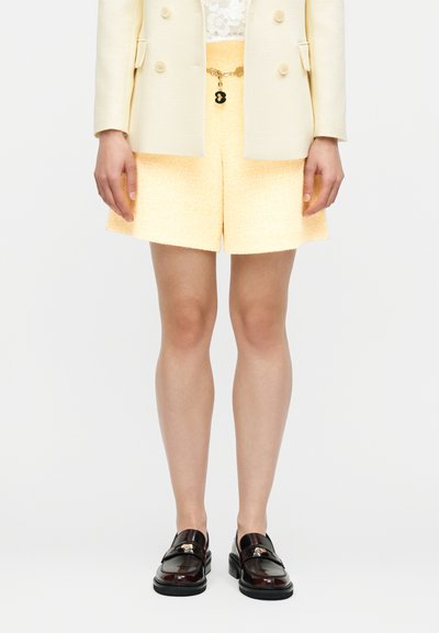 Personne portant un short texturé jaune clair avec un haut en dentelle blanche, un blazer crème et des mocassins marron foncé avec des accents dorés.