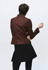 Persona con capelli corti vestita con un blazer marrone e una gonna nera, in piedi con le spalle alla macchina fotografica e un braccio piegato al gomito.