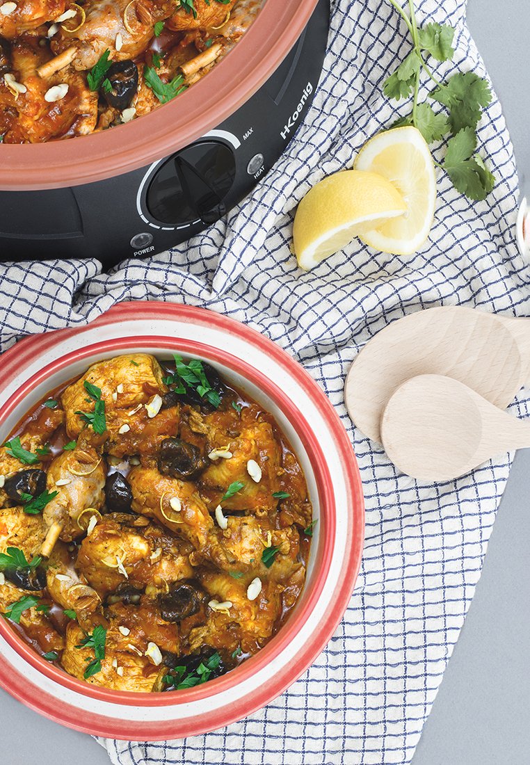 Rote und weiße Keramitschüssel mit Hühnersteak, garniert mit Petersilie, Mandeln und Oliven. Schwarzer Slow Cooker, Holzbesteck und Zitronenscheiben in der Nähe.