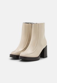 Stivaletti alla caviglia in vernice beige con tacco a blocco, caratterizzati da una superficie liscia e dettagli in cucitura, che si estendono sopra la caviglia per un look aderente.