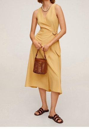 Kvinde iført en ærmeløs sennepsfarvet slå-om-kjole med spænde, holder en brun flettet håndtaske og har mørkebrune sandaler på.