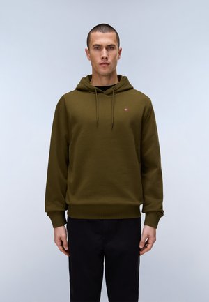 Man standing facing forward wearing an olive green hooded sweatshirt and black pants against a plain light background.