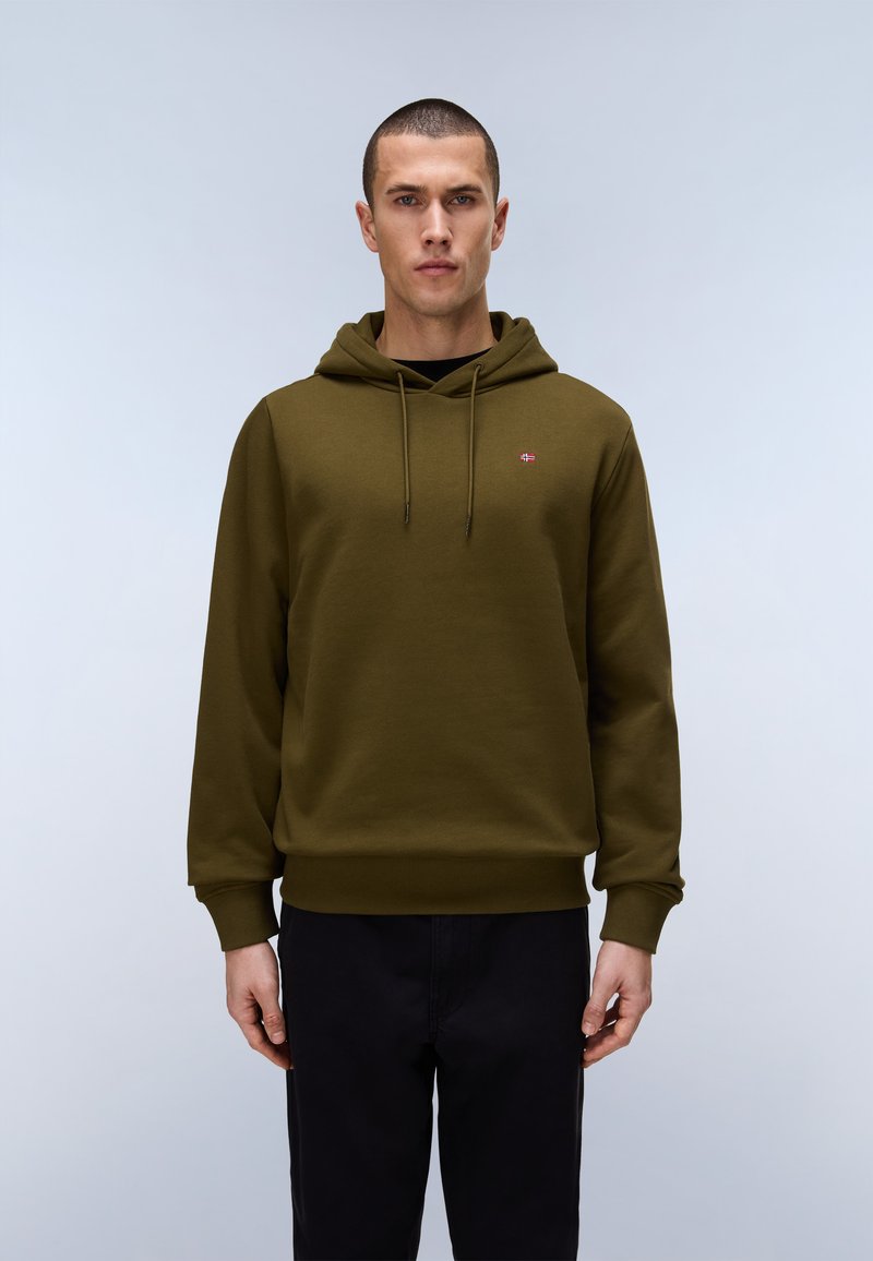 Muž stojící čelem vpřed, oblečený v olivově zelené mikině s kapucí a černých kalhotách na světlém jednobarevném pozadí.