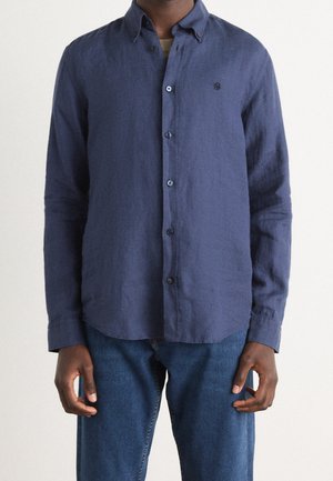 Mężczyzna w granatowej koszuli z długim rękawem zapinanej na guziki, noszący niebieskie jeansy, stoi z rozluźnionymi rękami wzdłuż ciała na jednolitym tle.