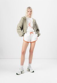 Sudadera gris clara con hombros caídos, camiseta blanca con un logo rojo, pantalones cortos blancos con ribetes naranjas y zapatillas blancas con detalles verdes.