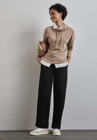 Felpa beige con cappuccio e cordoncino, indossata sopra una camicia bianca con colletto. Pantaloni neri a gamba larga con finitura strutturata, abbinati a sneakers bianche.