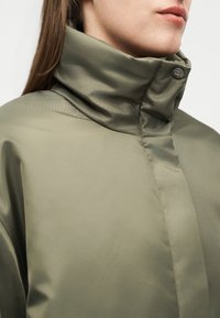 Γυναίκα που φοράει μπουφάν βαμβακερό ελιάς με ψηλό γιακά και κλειστή πατιλέτα με κουμπιά στον λαιμό, ορατή από τους ώμους μέχρι το πηγούνι.