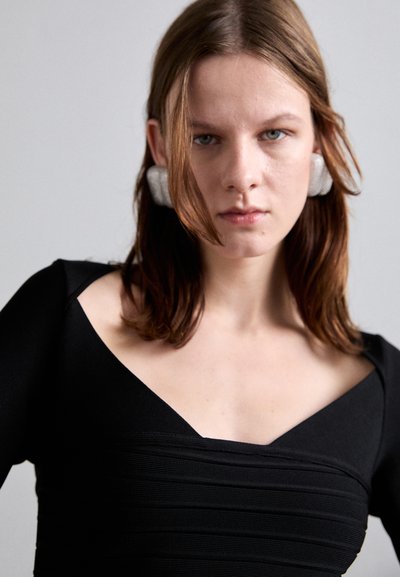 Svart, åtsittande klänning med strukturerad banddetaljering och bred rynkad halsringning. Modellen bär grå tygaccessoarer i håret. Mjuk, slät textur.