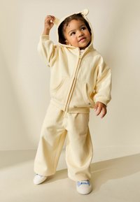 Roomkleurige fleece hoodie met berenoren, gecombineerd met wijde broekspijpen. Voorzien van zakken aan de voorkant en een ritssluiting, gedragen met sneakers met blauwe accenten.