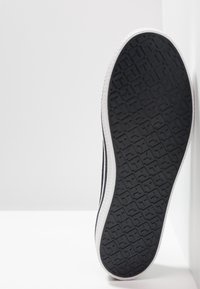 Semelle extérieure en caoutchouc d'une basket avec un motif de bande de roulement géométrique noir et une semelle intermédiaire blanche, positionnée à la verticale contre une surface blanche.