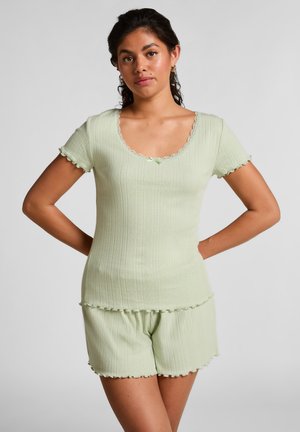 Woman in light green ribbed short-sleeve top and matching shorts, standing with hands behind back against plain background.