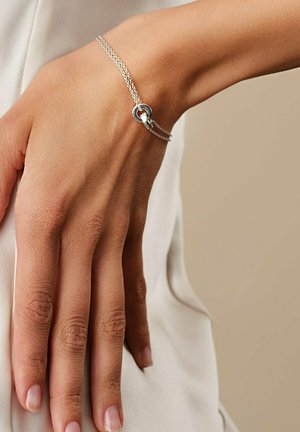 Hånd med et sølv dobbeltkædearmbånd med en rund vedhæng, hvilende på en hvid stofbaggrund.