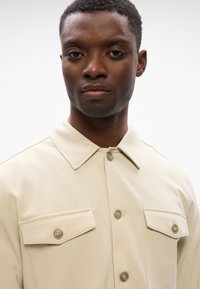 Μπεζ πουκάμισο με γιακά και λεία υφή, που διαθέτει δύο τσέπες στο στήθος και κουμπι ά μπροστά. Απλός σχεδιασμός χωρίς σχέδια ή διακοσμητικά στοιχεία.