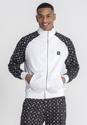 Rompevientos blanco con mangas de patrón negro, que cuenta con cierre de cremallera y puños elásticos. Pantalones a juego con el mismo patrón completan el conjunto.