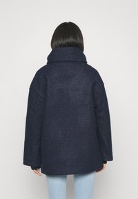 Mörkblå ullrock med hög krage, lös passform, texturerad yta och drop shoulders, sedd bakifrån. Ljusblå jeans i kombination.