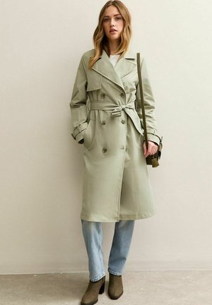 Mujer con un abrigo trench verde claro con cinturón, jeans azul claro y botines oliva, sosteniendo un bolso de hombro oliva.