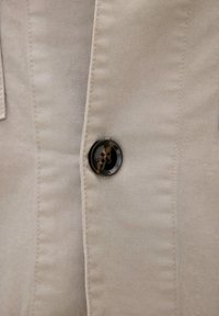 Close-up van beige stof met gedetailleerd stiksel en een enkele donkerbruine viergatenknop vastgemaakt op een kledingstuk.