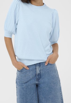 Mujer con top azul claro de manga corta abullonada metido dentro de unos vaqueros de mezclilla azul de cintura alta, con las manos en los bolsillos.