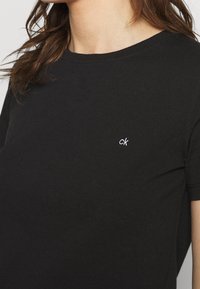 Zbliżenie na osobę ubraną w czarną koszulkę z okrągłym dekoltem, z małym białym logo "ck" na lewej piersi.