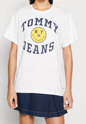 Witte katoenen t-shirt met marineblauwe tekst "TOMMY JEANS" en een gele smiley-logo. Gecombineerd met een donkere denim plooienrok.