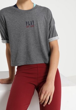 Person iført en grå cropped t-shirt med teksten "PLAY est. 2006" og højtaljede røde bukser, siddende med den ene hånd hvilende på en hvid overflade.