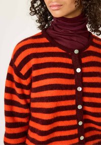 Cardigan rayé en orange et rouge foncé, fabriqué dans un matériau doux, avec un col en V et six boutons ressemblant à des perles sur le devant.