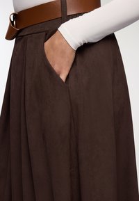 Mini gonna marrone in suede con tasca laterale, completata da una cintura in pelle marrone. Parte superiore bianca a maniche lunghe parzialmente visibile.