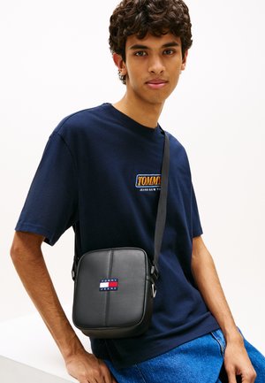 Schwarze synthetische Umhängetasche mit quadratischer Form, versehen mit einem bunten Logopatch und einem einfachen verstellbaren Riemen, getragen über einem marineblauen Hemd.