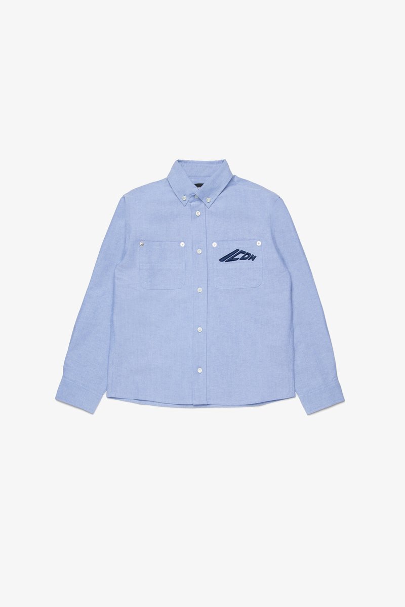 Light blue button-up shirt made of woven fabric, featuring two chest pockets and navy embroidered logo on the left side.