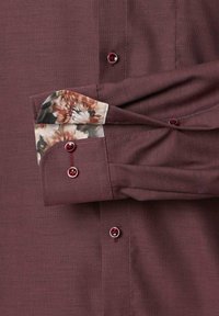 Burgunderfarbendes Hemd mit Knöpfen und strukturiertem Gewebe, das Blumenmuster an den Manschetten und passende burgunderfarbene Knöpfe entlang der Front aufweist.