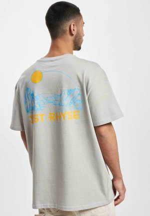 Lys grå bomulds t-shirt med et blåt og gult bjerglandskab grafisk design med sol og teksten "JST RHYSE" på bunden. Korte ærmer.