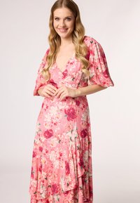 Robe fleurie rose avec de courtes manches bouffantes, un col en V et un ourlet à étages. Comprend des roses roses et des fleurs blanches, fabriquée en tissu léger.