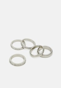 Sett med fem metallringer: fire glatte sølvband og én med klare krystaller langs kanten. Glatt finish, varierende bredder.