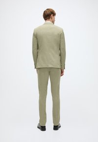 Ανοιχτό πράσινο λινό κοστούμι με στενή εφαρμογή, που διαθέτει γιακά με χαραγμένη εγκοπή και δύο κουμπιά στο σακάκι. Το παντελόνι ταιριάζει με το σακάκι στο χρώμα και την υφή.