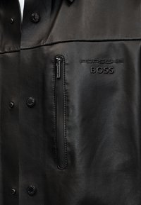 Μαύρο δερμάτινο μπουφάν με κάθετη τσέπη με φερμουάρ και ανάγλυφα λογότυπα "Porsche BOSS" στο στήθος, με κλείσιμο με κουμπιά πατιλέτας.