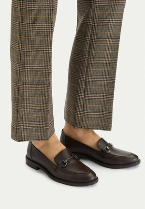 Person trägt braune Leder-Loafer mit silberner Metallschnalle und karierte, gekürzte Hosen vor einfarbigem Hintergrund.