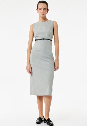 Kobieta w bezrękawowej, szarej sukience do kolan z cienkim czarnym paskiem i czarnymi mokasynami, stojąca na tle białego, gładkiego tła.