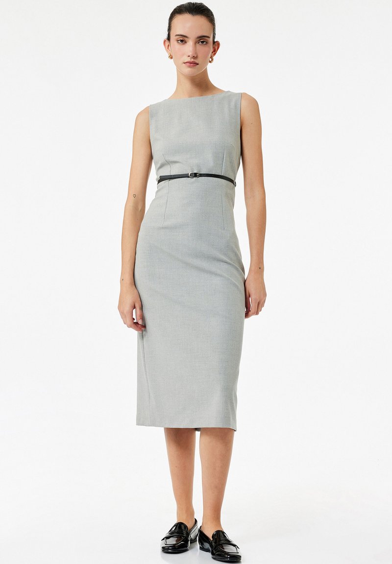 Kobieta w bezrękawowej, szarej sukience do kolan z cienkim czarnym paskiem i czarnymi mokasynami, stojąca na tle białego, gładkiego tła.