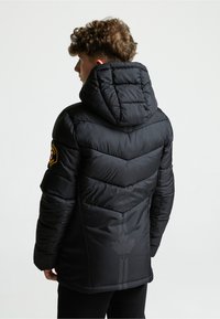 Melnā piedurkņu jaka ar kapuci, ar ševronu šuvēm un teksturētu aizmuguri, kas rotāta ar atstarojošu rakstu un logo uz piedurknes.