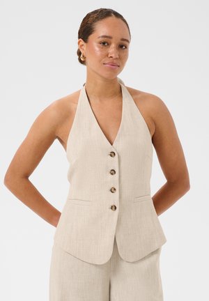 Femme aux cheveux tressés portant un gilet beige sans manches boutonné et un pantalon assorti, debout les mains dans le dos sur fond blanc.