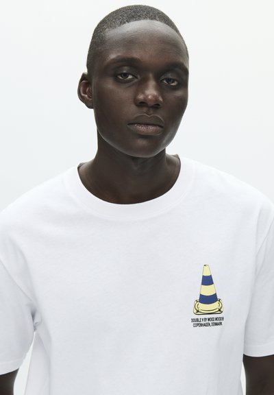 Λευκό βαμβακερό t-shirt με πολύχρωμο γραφικό ενός κώνοντας κυκλοφορίας στο αριστερό στήθος, με μπλε και κίτρινες ρίγες και κείμενο της εταιρείας από κάτω.