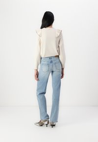 Suéter de crema con acentos de volantes en los hombros, combinado con jeans rectos de color azul claro y zapatos de tacón de metal plateado. Fondo minimalista.