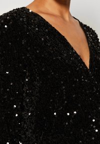 Svart paljetttyg med en texturerad yta, med en djup V-ringning och skimrande accenter från paljetterna genom hela tyget.