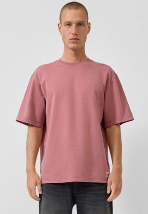 Mężczyzna stojący przodem, ubrany w luźną, różowawą, krótko rękawkową koszulę i czarne spodnie na jednolitym jasnym tle.